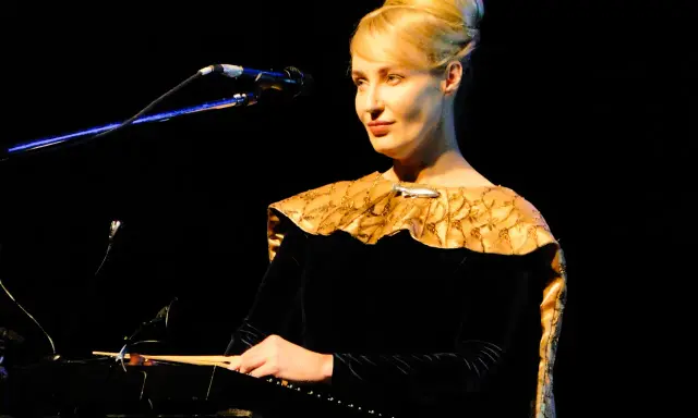 Kobieta w czarnej sukni z ozdobnym kołnierzem gra na instrumencie, tworząc muzykę. Kto śpiewa?