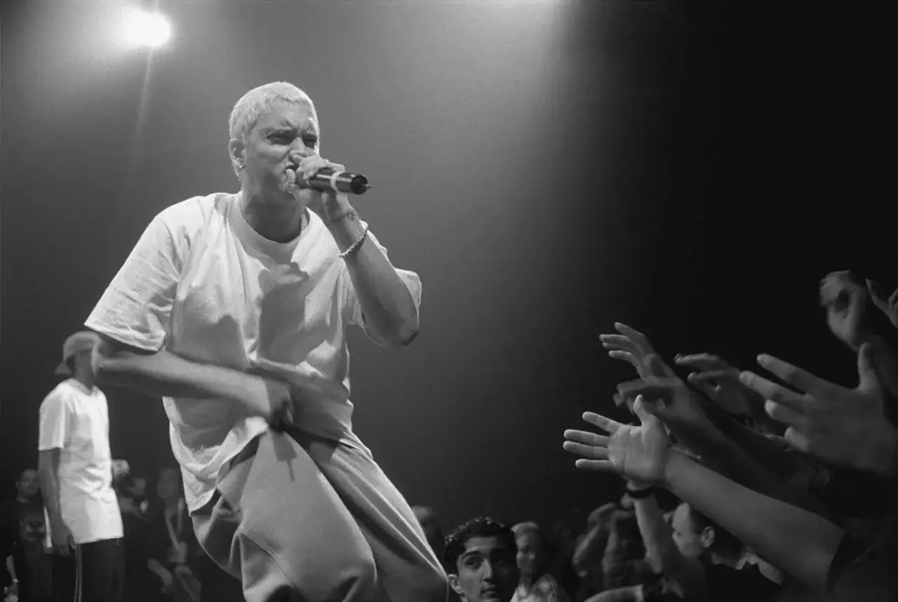 Eminem na scenie, w białej koszulce, śpiewa do mikrofonu, a tłum wyciąga ręce. Tak właśnie dzieje się z Eminemem podczas koncertu.