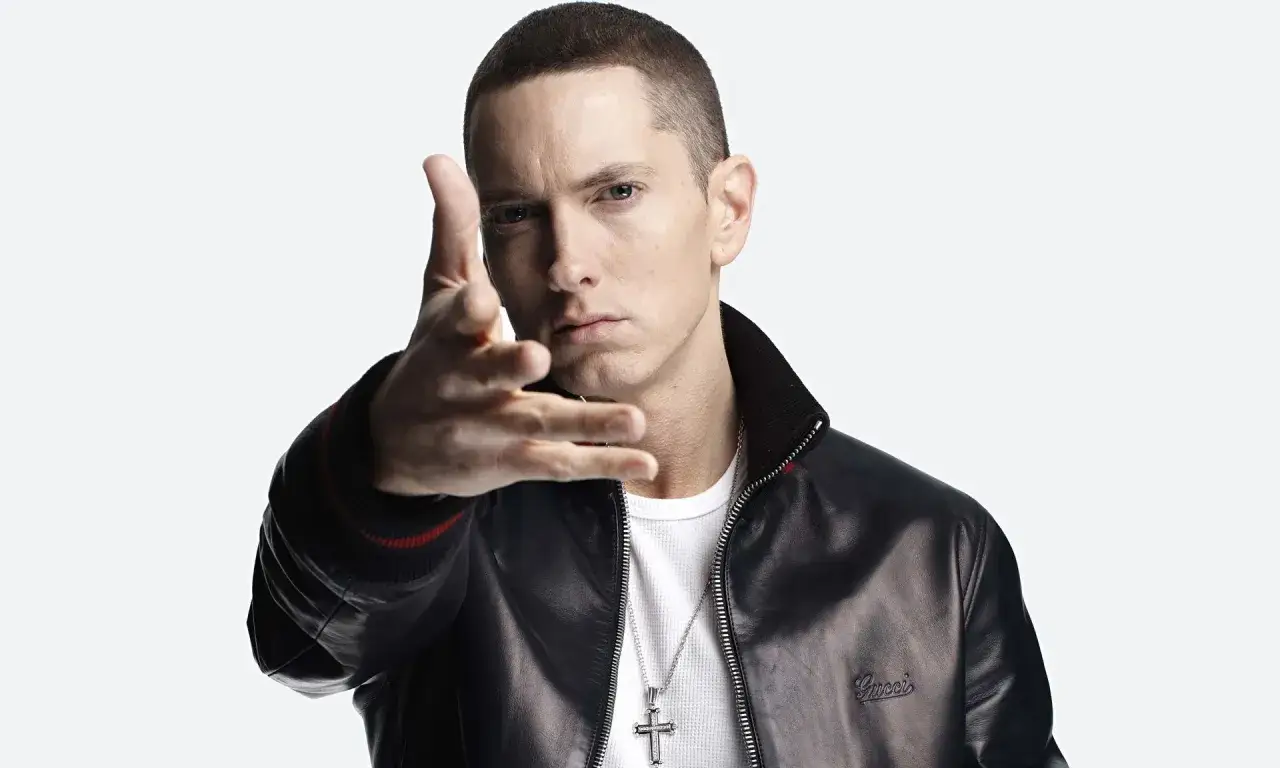 Eminem w czarnej kurtce i białej koszulce, z krzyżem na szyi, wyciąga rękę. Choć nie widać tego na zdjęciu, wiele os&oacute;b zastanawia się, ile Eminem ma wzrostu.