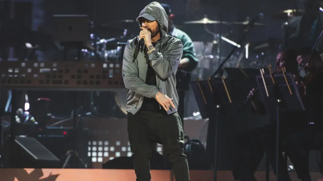 Eminem w szarej bluzie z kapturem i czapce rapuje na scenie, podczas gdy orkiestra gra w tle. Tak właśnie dzieje się z Eminemem na koncercie.
