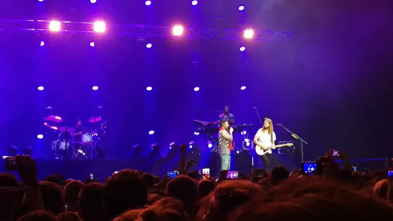 Koncert Maroon 5 w pełnym blasku fioletowych świateł. Fani nagrywają występ, zastanawiając się, ile trwa koncert Maroon 5.