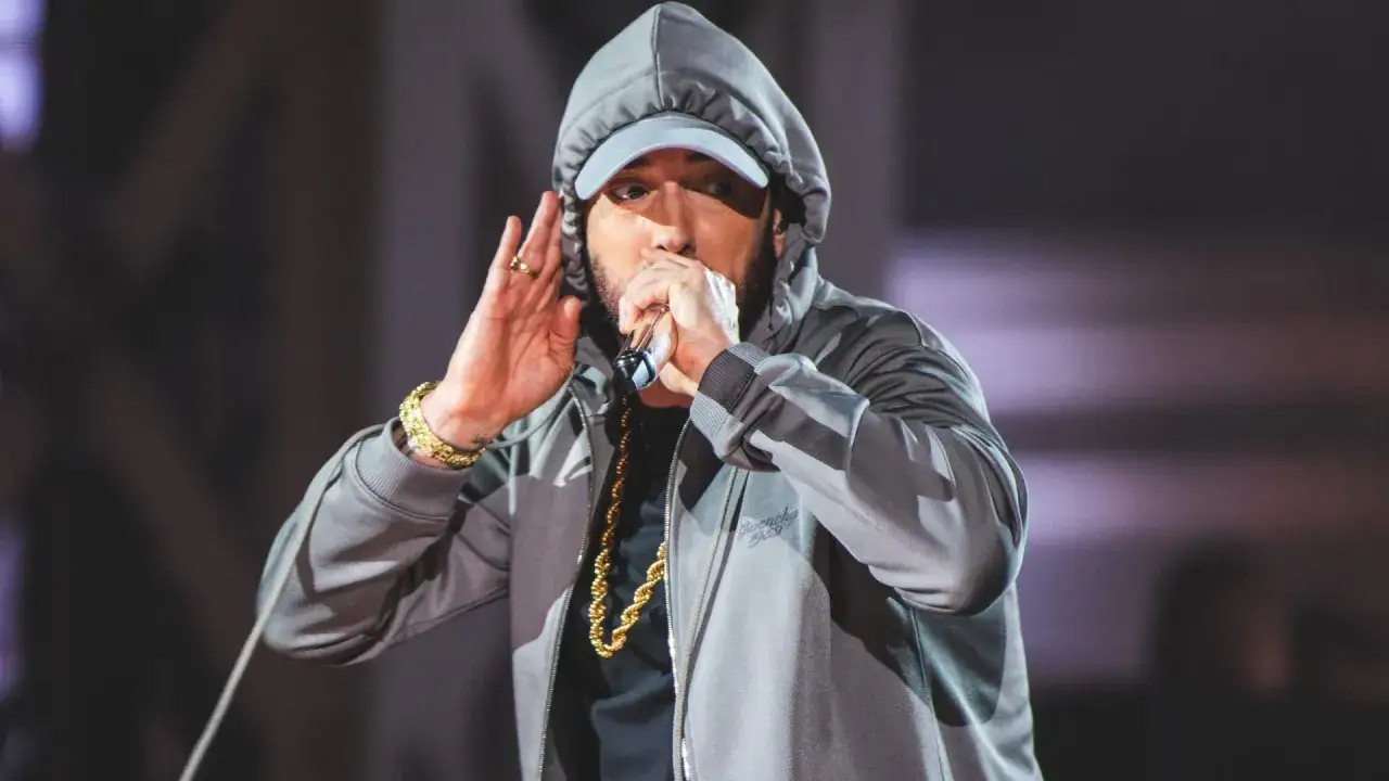 Eminem w szarej bluzie z kapturem i czapce, z mikrofonem w dłoni, słucha publiczności. Co się dzieje z Eminemem? Koncert trwa!