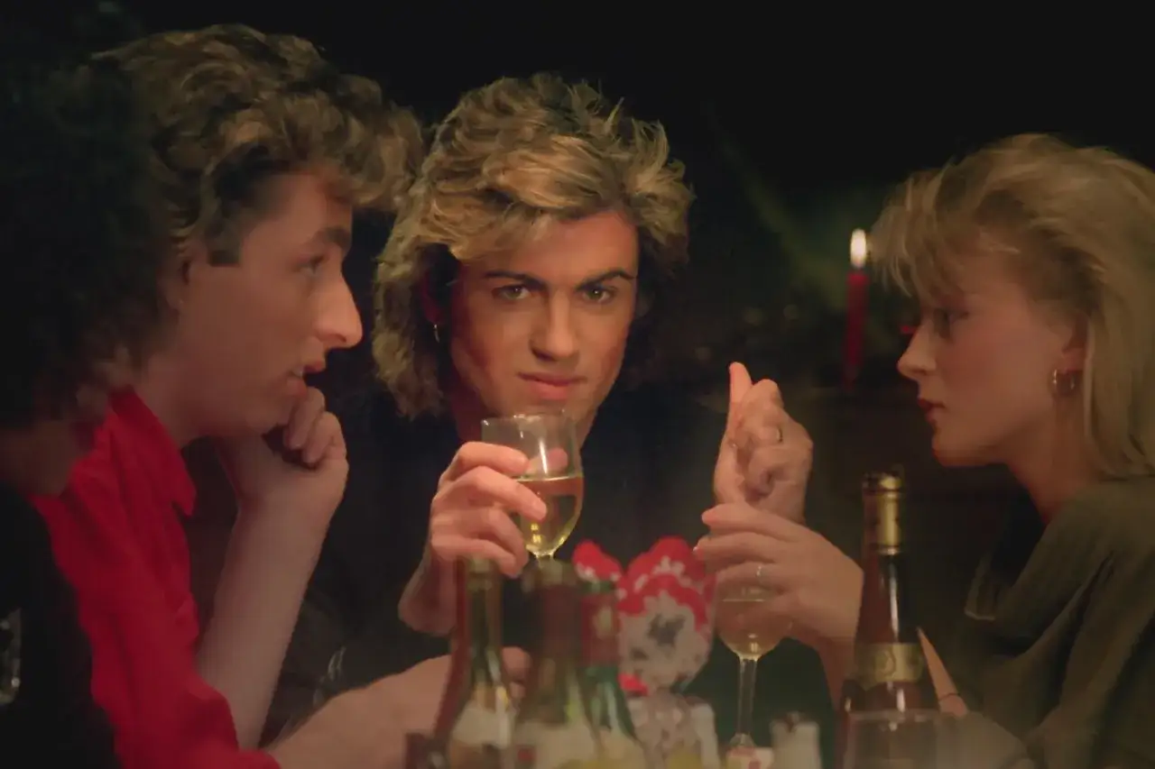 George Michael, kt&oacute;ry śpiewa Last Christmas, siedzi przy stole z przyjaci&oacute;łmi, pijąc wino.