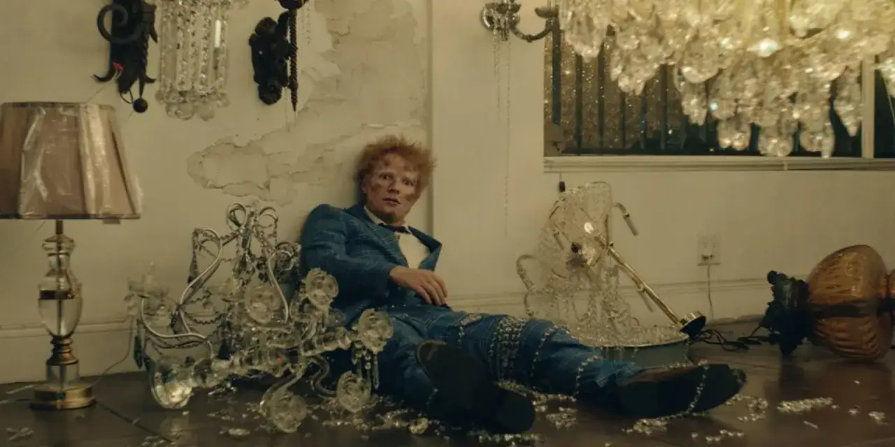 Ed Sheeran siedzi pośród zniszczonych kryształowych żyrandoli i lampy. Wygląda, jakby przeżył katastrofę, może to był dzień, kiedy urodził się Ed Sheeran.